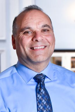 A portrait of Corey Farris who is smiling while wearing a blue shirt and blue patterned tie.