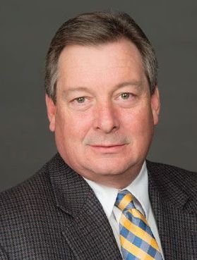 This is a portrait of Ron Justice. He is sitting in front of a dark gray background, has short dark hair and is wearing a dark plaid jacket and gold and blue plaid tie with a white button-up shirt.