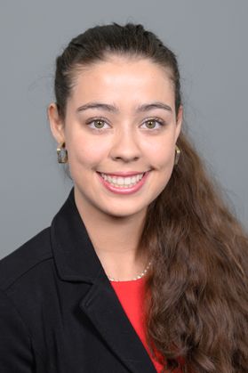 Headshot of WVU Bucklew Scholar Anna Brusoe. She is pictured against a gray background and is wearing a black jacket over a red blouse. She has long, wavy brown hair and is wearing gold hoop earrings. 