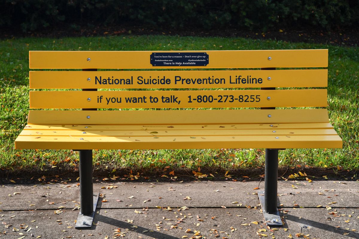 Image showing an outdoor yellow bench on a sidewalk. There is green grass behind it. The toll free number for the National Suicide Prevention Lifeline is printed on the bench in black letters. 