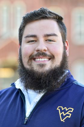 A man with dark hair and a dark beard wearing a blue WV zip-up top poses for a headshot.