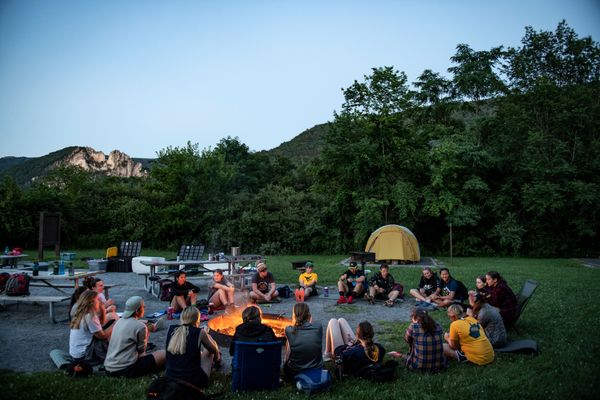Photograph of WVU students sitting around a glowing campfire. Dusk is setting in, a yellow tent is off in the distance as well as various camping gear. The Seneca Rocks formation is on the horizon above them. 