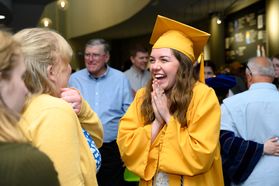 A graduate in a gold cap and gown is shown in the lobby of the Creative Arts Center clapping her hands. A crowd of family and friends surrounds her.
