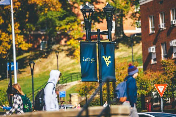 Students walking on campus with backpacks on. They are wearing hoodies with hats on, and the leaves of the tress are beginning to turn colors. A WVU 
