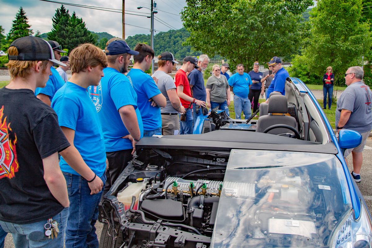 A large group of people looking at an electric vehicle. Many of them are in blue shirts.