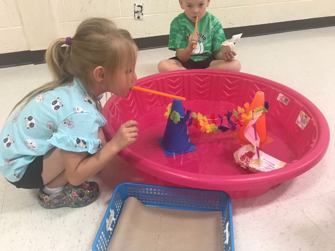 Young children participating in an educational game. One is wearing a blue shirt and has a long blond ponytail. the other is wearing a green shirt. they are sitting next to a small pink plastic pool with no water. Inside the pool is a game.
