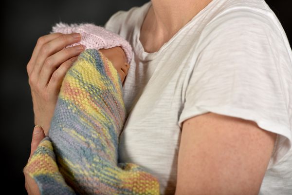 An image of a female, from the neck down, wearing a white t-shirt holding an infant wrapped in a multi-colored blanket and wearing a pink knitted hat. 