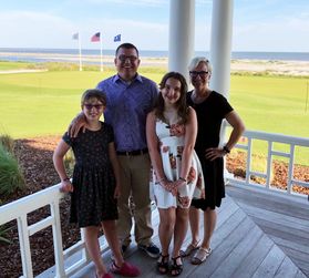 Picture of the Alban family, recent donors to a new STEM scholarship at WVU. Bob Alban is standing outside with his wife and two daughters. There is a beach in the distance and expanse of green grass beyond them. 