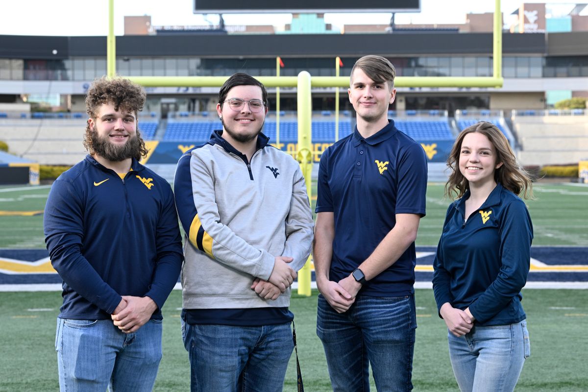 The four finalists to be the 69th Mountaineer mascot stand at the end zone on Mountaineer field. The four are all wearing tops with the Flying WV on their left shoulders and they are standing with their arms crossed.