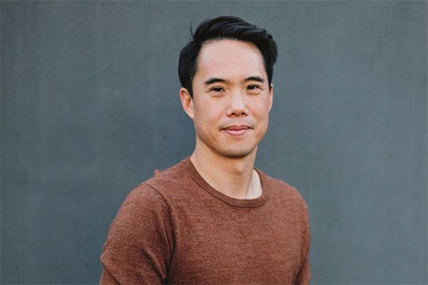 Author Charles Yu is pictured here. He is of Asian descent and is wearing a rust colored sweater. he has short, black hair. 