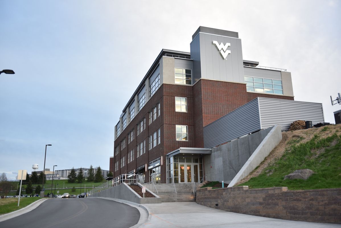 A building with grey and brick walls and a WVU logo. The sky is blue with white foggy clouds.