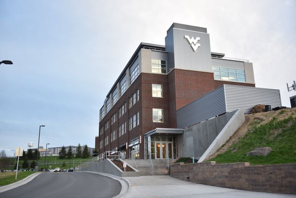 A building with grey and brick walls and a WVU logo. The sky is blue with white foggy clouds.