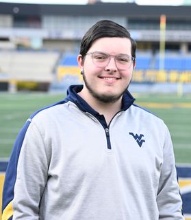 This is a portrait of Christian Adkins. Christian is wearing a gray WVU pullover with a dark blue Flying WV on the left chest and is standing on Mountaineer Field.