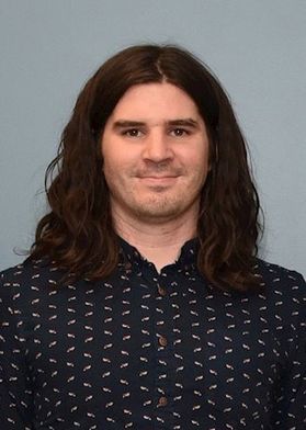 Photograph of WVU alumnus Michael Morehead. He is pictured against a light gray background wearing a black and white patterned buttoned up shirt. He has long brown hair.