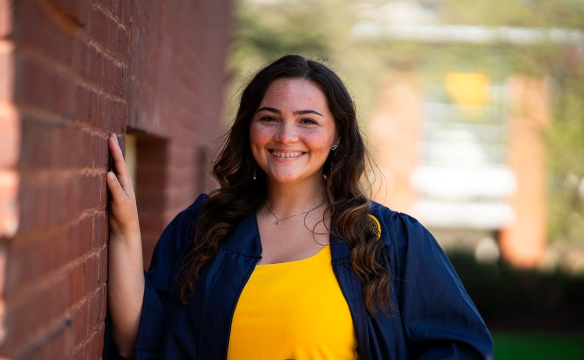 Photograph o WVU School of Dentistry student Mackenzie Wensel wearing a blue gown over a gold blouse. She has long, brown hair and is standing against a red, brick wall. 