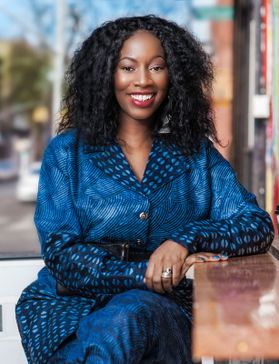 Headshot of WVU honoree Farai Simoyi. She is seated outside at a table wearing a bright blue subtly patterned dress. Her dark hair is long and curly. 
