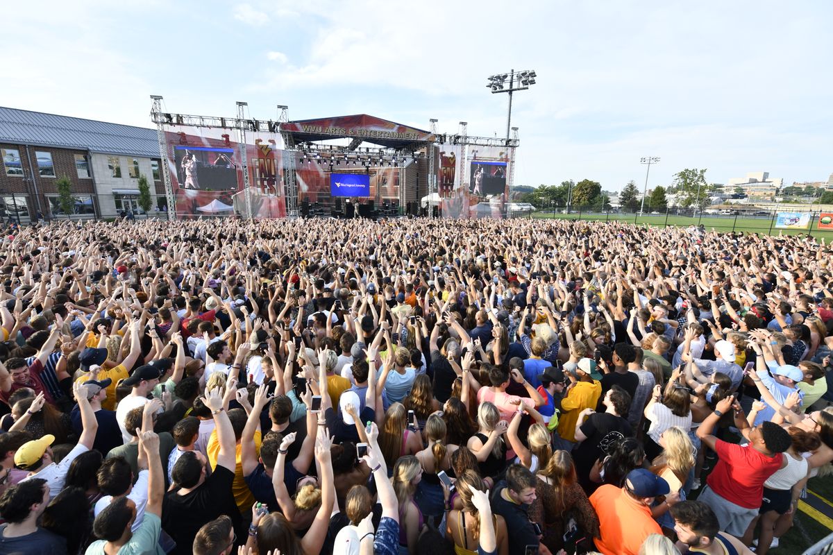A performance stage is in the back of this photo. Between it and the camera are hundreds of people facing the stage with their hands in the air.