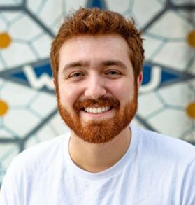 This is a portrait of Travis Rawson who has red hair, a red beard and is smiling while wearing a white T-shirt in front of a gold and blue WVU tiled mosaic.