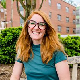 Headshot of WVU employee Olivia Dale Pape. She is pictured outside with green hedges behind her. She is wearing an emerald green short sleeved shirt and has long, red hair. She also wears glasses. 