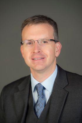 Headshot of WVU Professor Erik Herron. He is pictured against a gray background wearing a dark gray three-piece suit with a light blue dress shirt and a slate blue colored tie. He has short graying hair and wears frameless glasses. 
