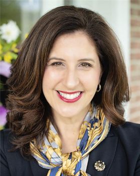 Headshot of WVU Professor Lisa DeFrank-Cole. She is piuctured outside wearing a colorful silk scarf tied around her neck. She has long brown hair and is wearing red lipstick. There are flowers and a brick wall behind her. 
