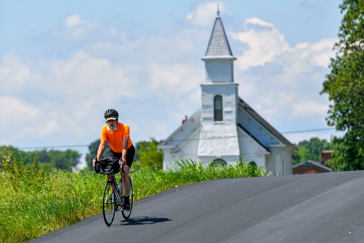 A person wearing a bright orange t-shirt riding a bike near a white church. The sky is blue and there is grass along the paved road.