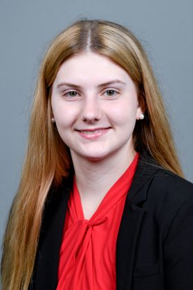 Headshot of WVU Bucklew Scholar Juliana Veazey. She is pictured against a gray background wearing a black jacket over a red blouse. She has long blonde hair. 