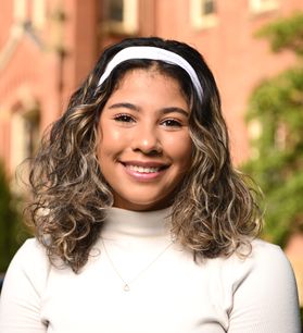 Headshot of WVU scholarship recipient Ariana Burks. She is pictured outside with Woodburn Hall in the background. She is wearing a cream colored turtleneck and matching headband. She has shoulder length brown hair with light colored highlights. 