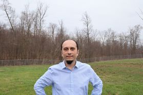 Man stands with hands on hips wearing a light blue shirt in a grassy field