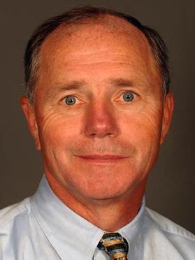 Headshot of John Spiker. He is pictured against a gray background and is wearing a light blue dress shirt. He has gray short hair and light colored eyes. 
