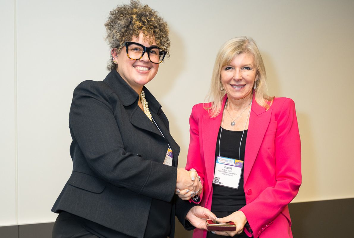 WVU faculty member Eloise Elliot receives a fellowship award from another woman dressed in black. Elliot is wearing a hot pink jacket and has shoulder length blonde hair. 