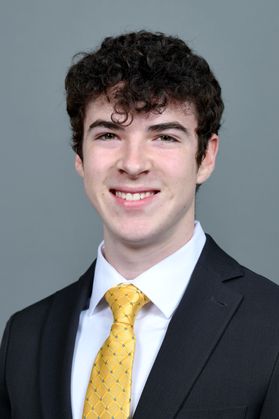 Headshot of WVU Foundation Scholar Joss Poteet. He is pictured against a gray background wearing a black suit over a white dress shirt and a yellow, patterned necktie. He has short, curly black hair. 