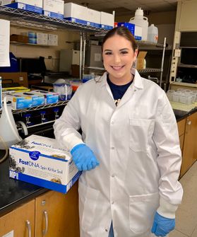 Photograph of WVU student Kara Cunningham. She is pictured in a lab wearing a white lab coat and blue safety gloves. Her brown hair is pulled back. 