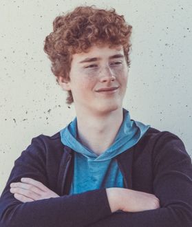 This is a portrait of Daniel Curtis who has short curly red hair and is wearing a blue shirt under a darker hoodie.