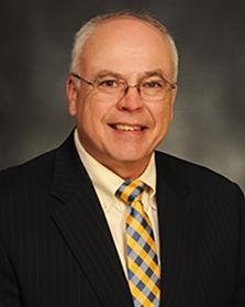 Headshot of WVU Health Sciences administrator Dr. Bill Ramsey. He is pictured in front of a dark gray muddled background and is wearing a dark colored suit over a yellow dress shirt with a blue and gold checkered tie. He has short gray hair and glasses.