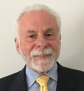 Sociology professor Henry Brownstein. He is posing for a headshot and has white hair, a white beard, and is wearing a dark coat, light blue shirt and yellow tie. 