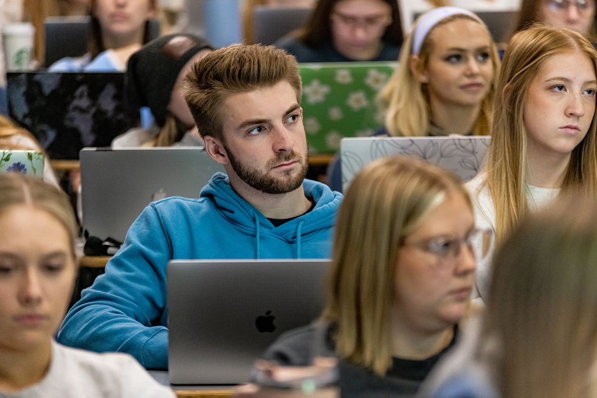 A man with raised hair and a beard sits in a classroom surrounded by peers with a laptop in front of him