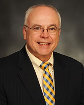 Headshot of WVU Medicine executive Bill Ramsey. He is pictured against a gray background and is wearing a dark colored suit with a pale yellow dress shirt and yellow and blue checkered tie. He has short gray hair and wearing glasses. 