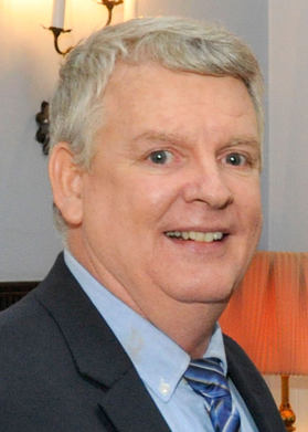 A person with short gray hair looks at the camera while wearing a dark suit jacket, blue button up shirt and striped blue tie.