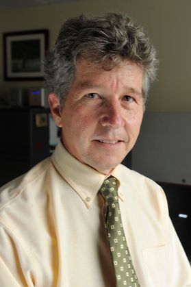 Headshot of WVU professor L. Christopher Plein. He is pictured standing in front of a bookshelf wearing a light-colored sport coat and an orange tie. He has salt and pepper hair and wears square framed glasses. His hands are causally clasped together.