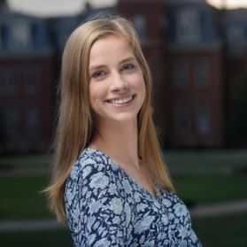 Headshot of WVU student Colleen Christopher. She s pictured outside with iconic Woodburn Hall behind her. She is wearing a blue floral top and has long, blonde hair. 