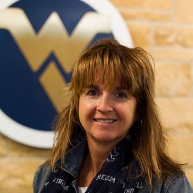 WVU professor Christine Schimmel. She is pictured against a tan wall with a circular Flying WV hanging on the wall. She has shoulder length light brown hair with bangs. She is wearing a denim top with a WVU scarf. 