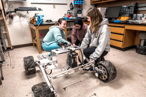 Three people are shown working on a robotics body with four black wheels on it in a lab. One person is in a gray sweatshirt with WVU on it.