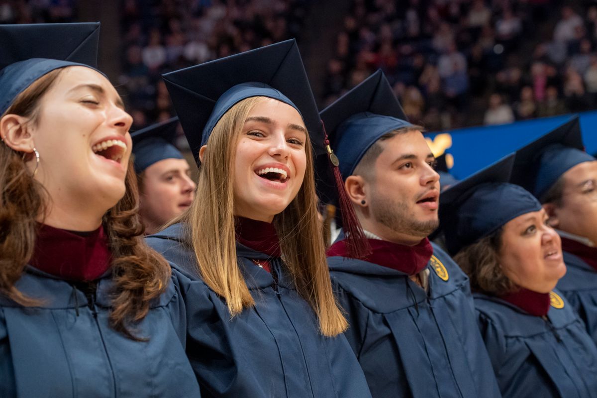 WVU graduates singing with their arms around each other at the commencement ceremony in their blue caps and gowns.