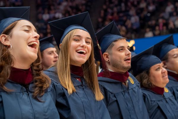 WVU graduates singing with their arms around each other at the commencement ceremony in their blue caps and gowns.