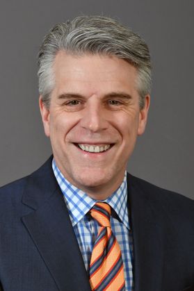 Photo of a smiling man wearing a blue and white checked shirt and orange and blue striped tie under a dark suit coat