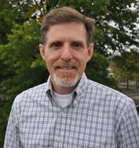 Headshot of WVU researcher Greg Dahle. He is pictured outside with green trees in the background. He is wearing a purple and while plaid shirt and has light colored hair with a gray gotee. 