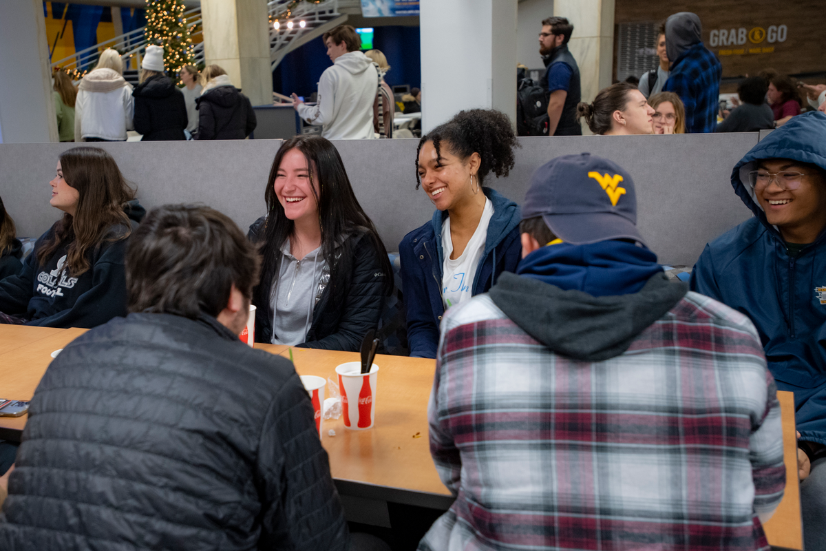 More than half a dozen students sit at a table in the Mountainlair. Two students have their backs to the cameras. Four others are facing them while leaning against a gray wall. Other students are walking beyond the wall.