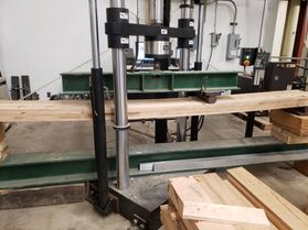 A large metal machine with black bars at the top and bottom presses down on a green bar stretching a couple of feet long. Underneath the green bar is a long wooden panel. This is a stress test for the wooden panel.