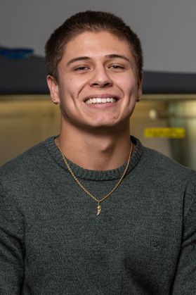 WVU student Christopher Smith poses for a headshot wearing a green sweater and a gold chain around his neck. He has very short dark colored hair and a wide smile. 